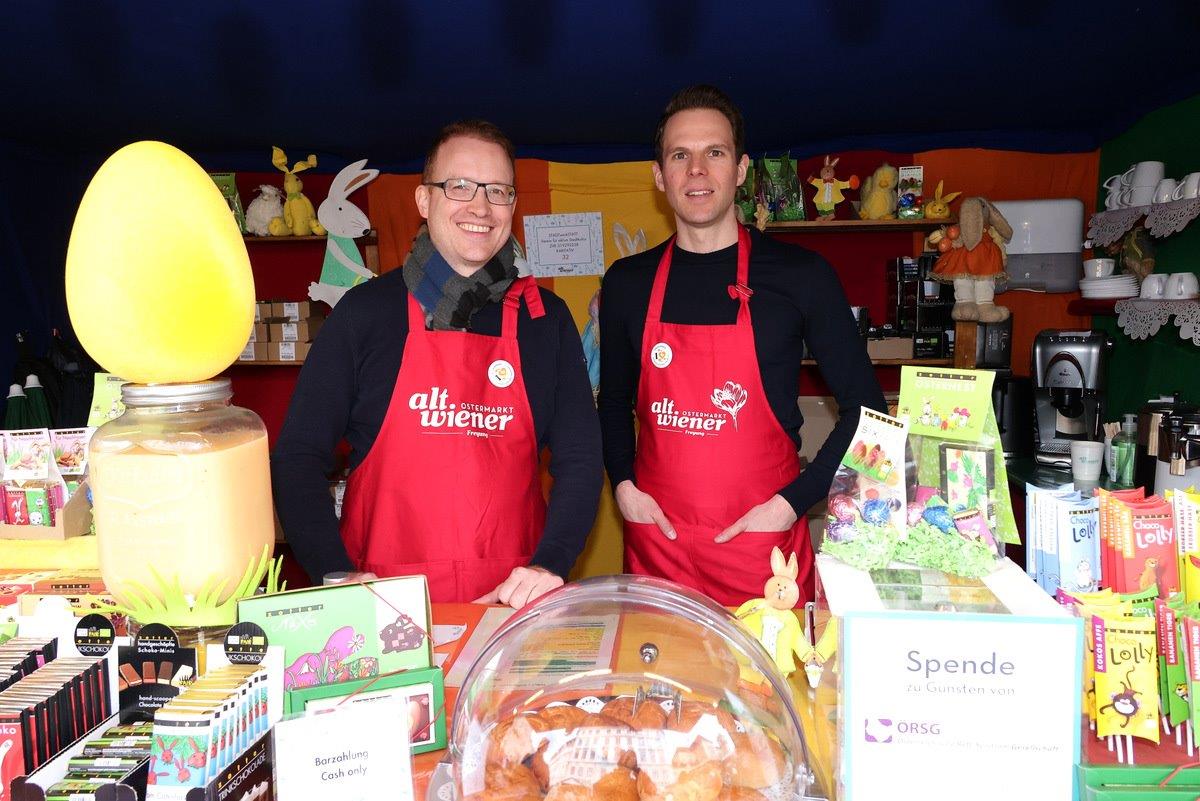 Two people in red aprons stand behind a colorful Easter-themed stall with chocolate, pastries, and decorations on display.