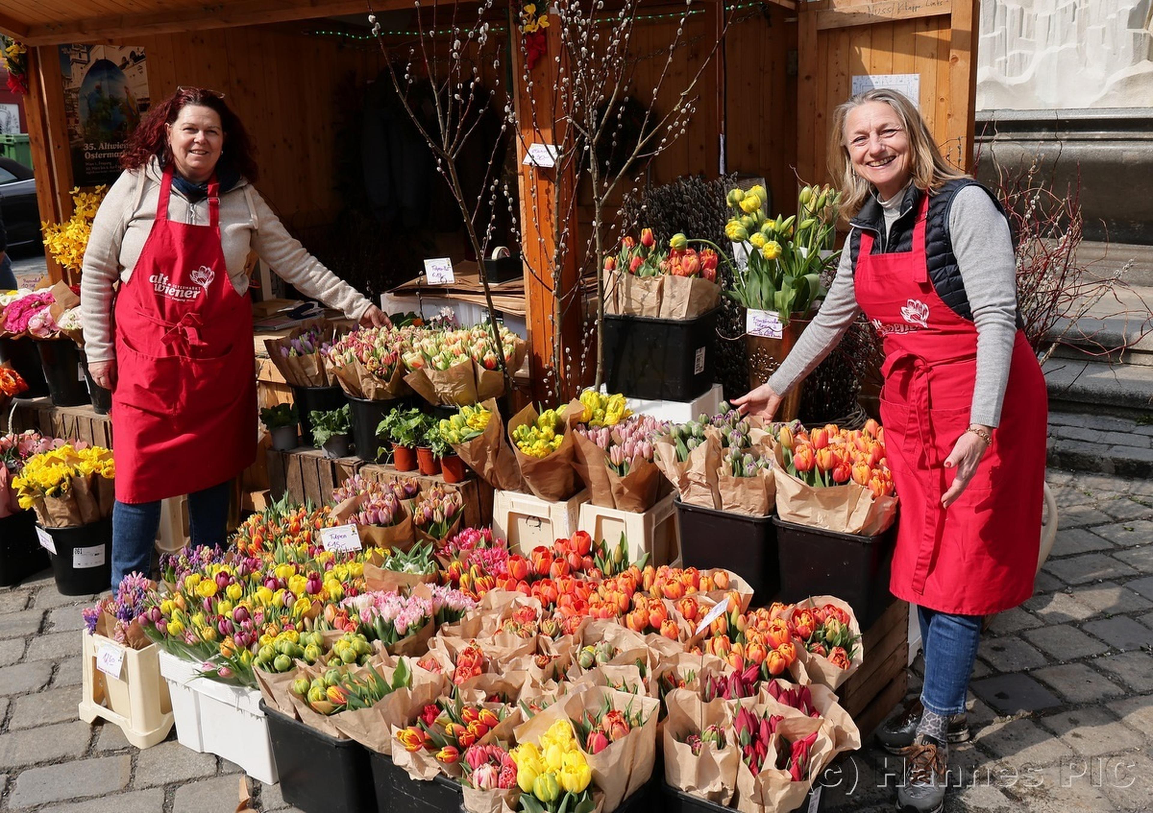 Two smiling vendors in red aprons display a variety of vibrant flowers, including tulips, at a market stall on a sunny day.