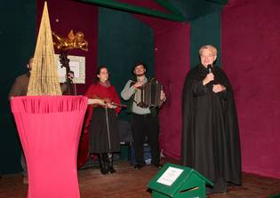 A person in a black robe speaks into a microphone, accompanied by musicians with a violin, accordion, and double bass, on a stage with red and green decor.