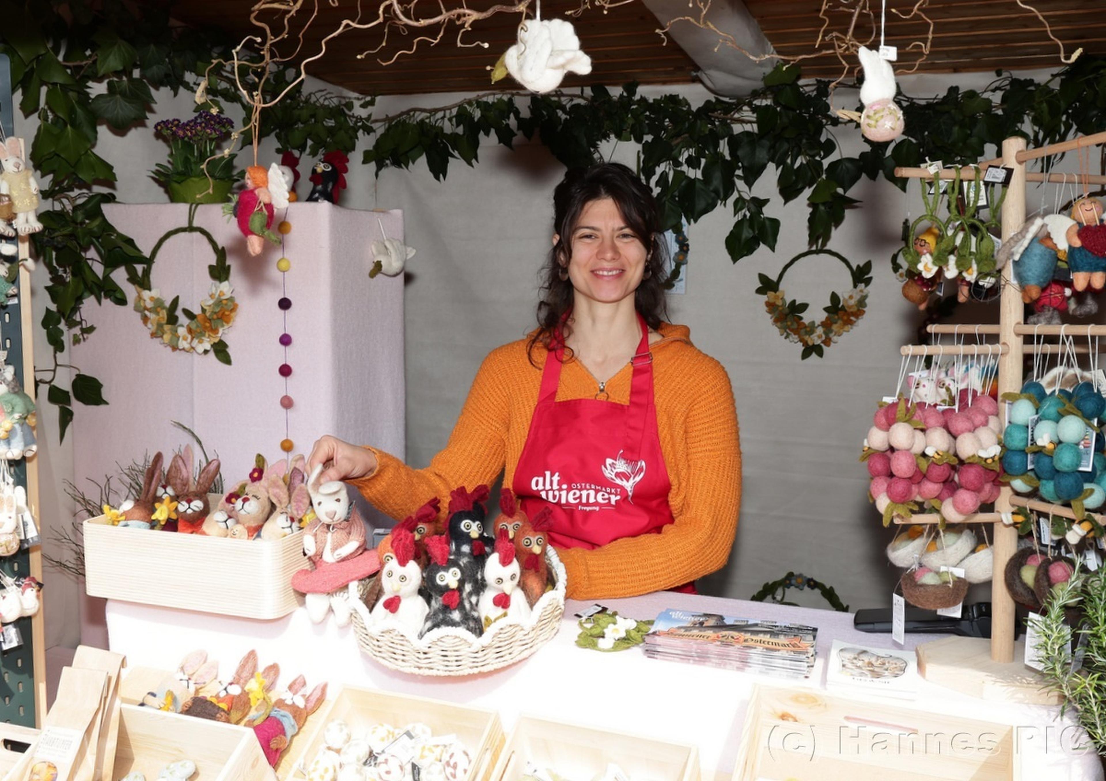 A woman in an orange sweater and red apron stands at a craft stall displaying handmade felt animals and decorations.