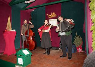 Three musicians perform on a small stage: one plays a double bass, another a violin, and the third an accordion. The stage is decorated festively.