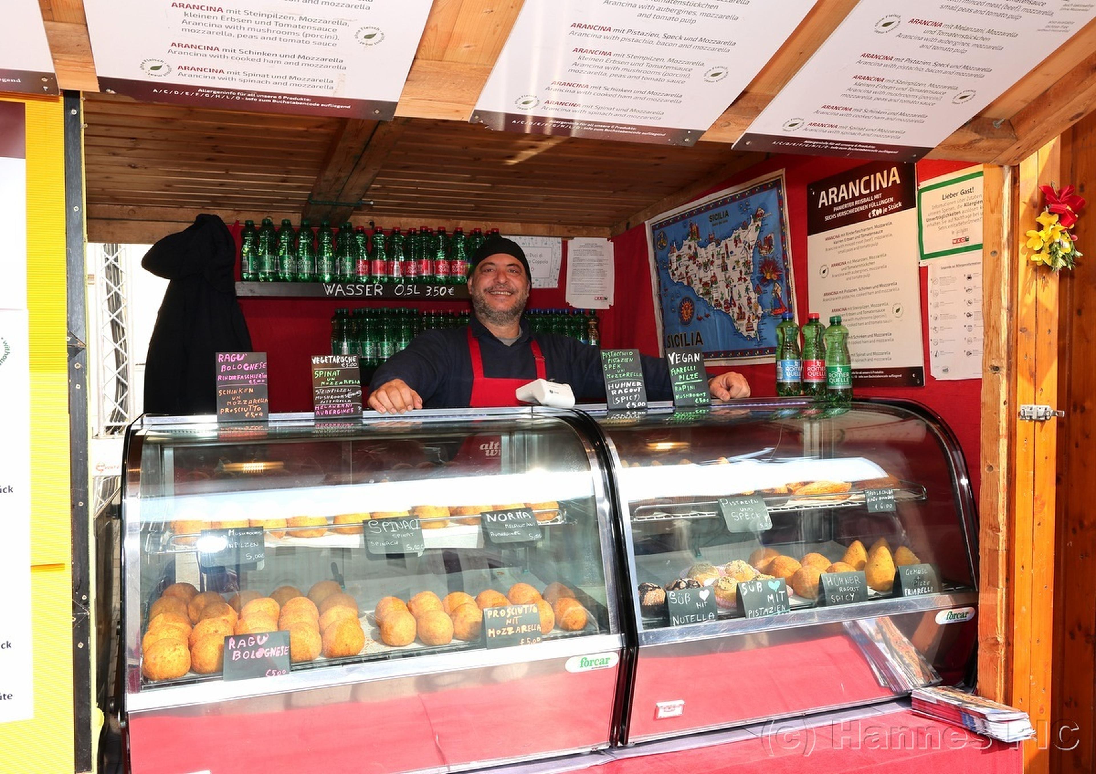 Man standing in a colorful food stall, displaying a variety of pastries and drinks, with a happy expression.