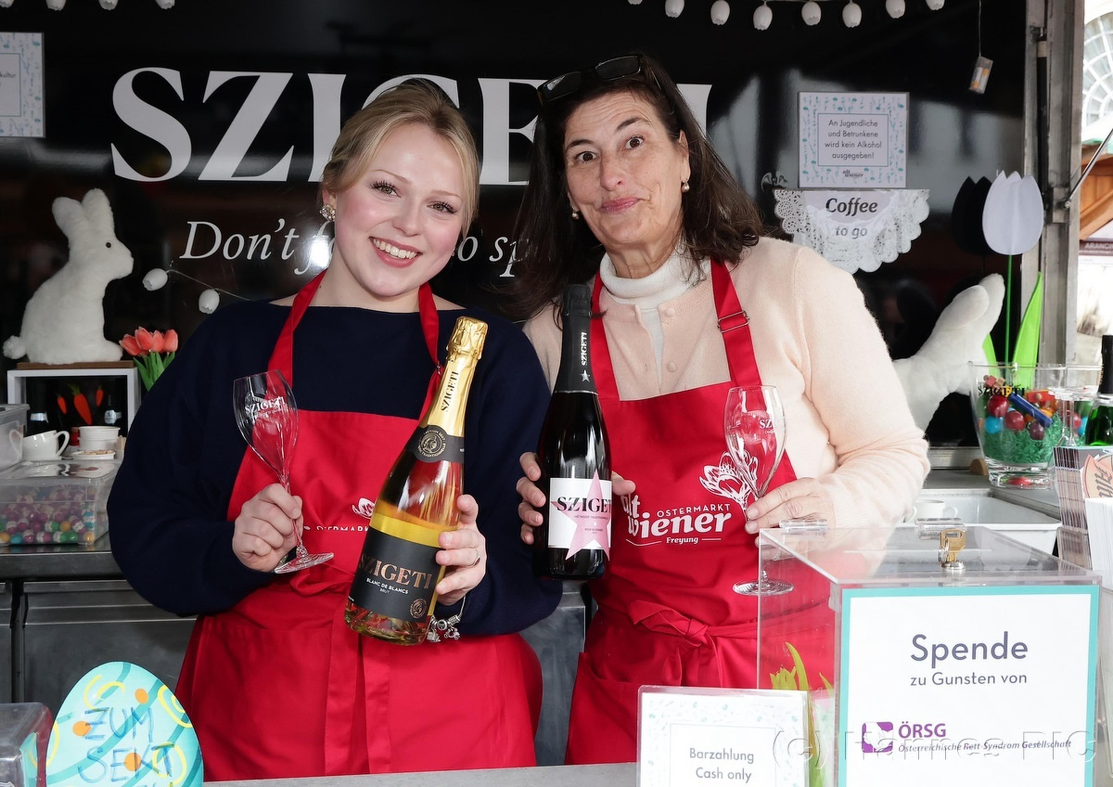 Two smiling women in red aprons hold bottles of wine at a booth, surrounded by glasses and donation signs.