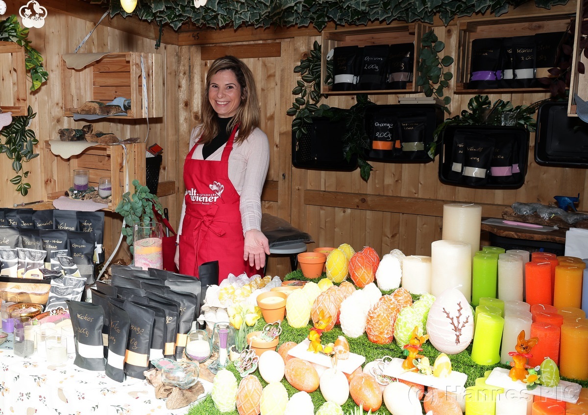 Woman in a red apron stands behind a wooden stall displaying colorful candles and decorative items, surrounded by greenery.