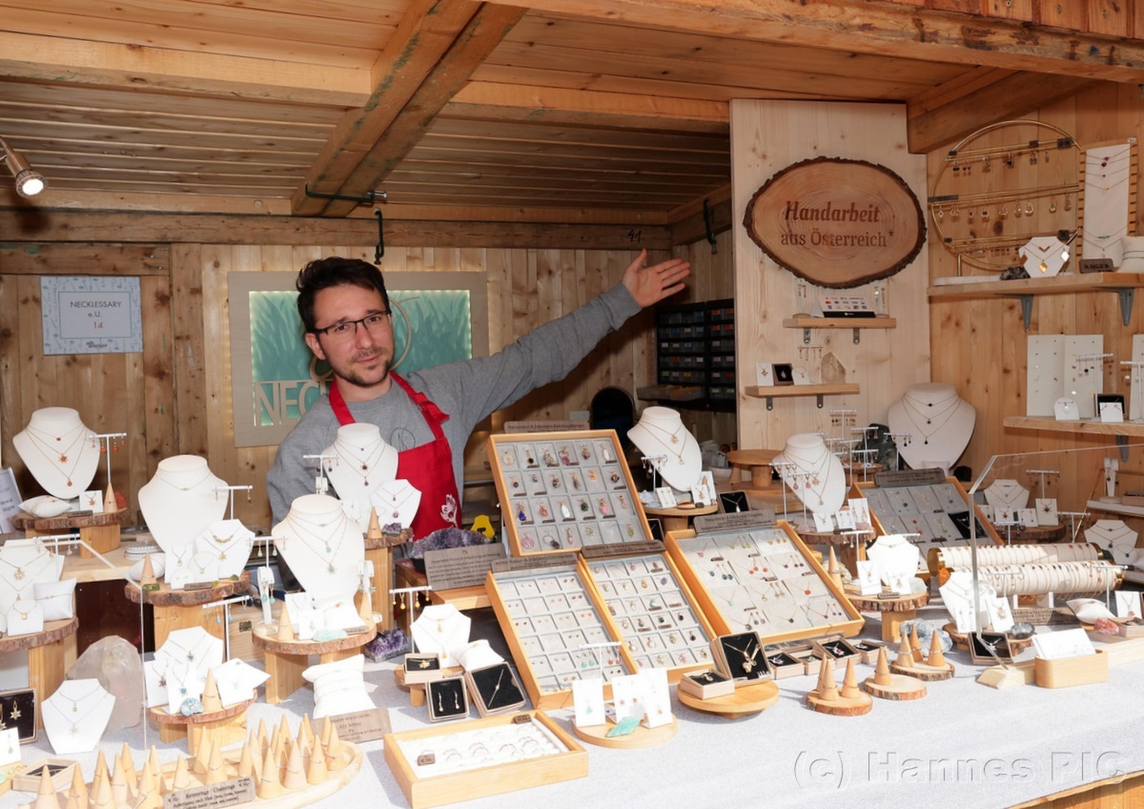 Man in a red apron presenting various jewelry displays in a wooden stall, featuring necklaces, earrings, and rings.