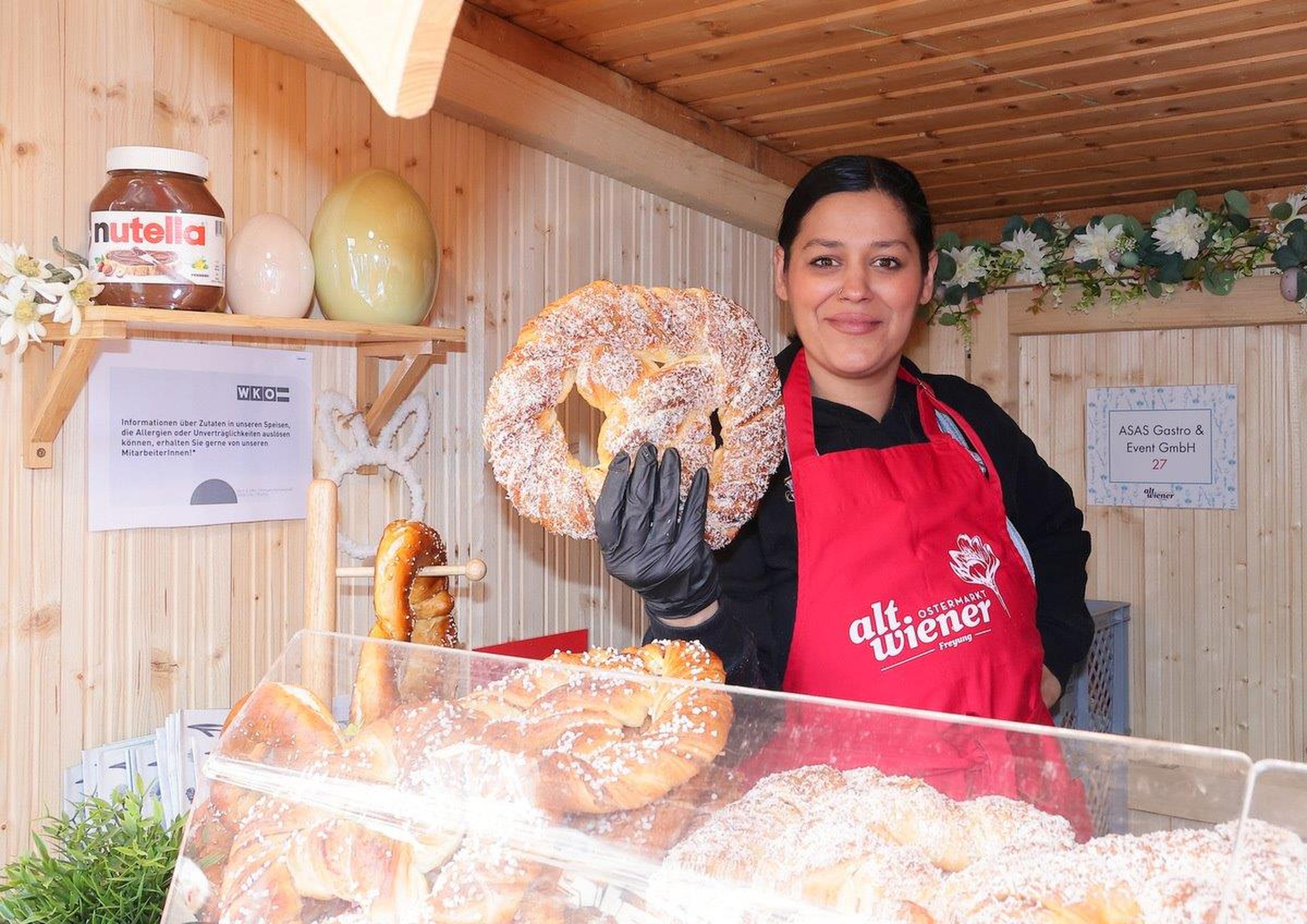 Smiling vendor holding a large pretzel, wearing a red apron. Various pretzels displayed in a wooden booth with Nutella jar in the background.