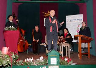 A man speaks passionately on stage, surrounded by musicians with guitars. The scene is festive, with candles, wreaths, and a banner reading "pro rare austria."