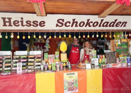 A festive market stall labeled "Heisse Schokoladen" displays chocolates and Easter items, with two people standing behind the counter.