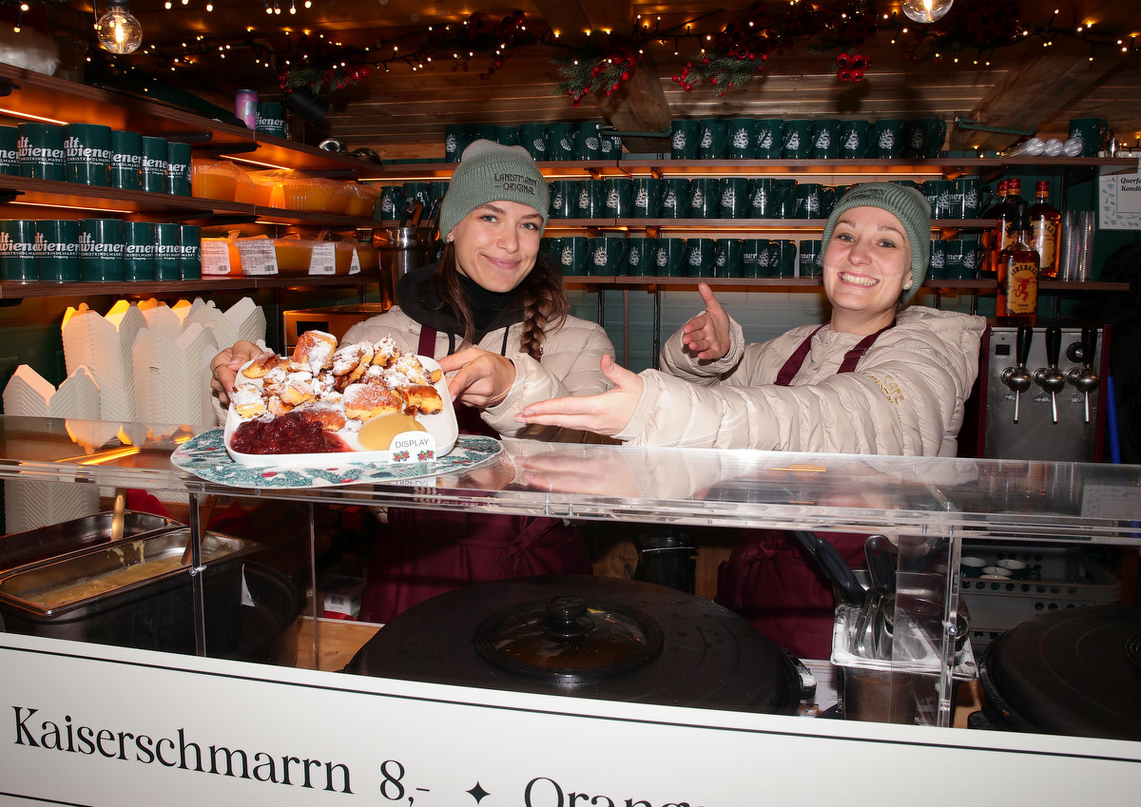 Two smiling vendors at a festive market stall present a plate of Kaiserschmarrn, surrounded by holiday decorations and lights.