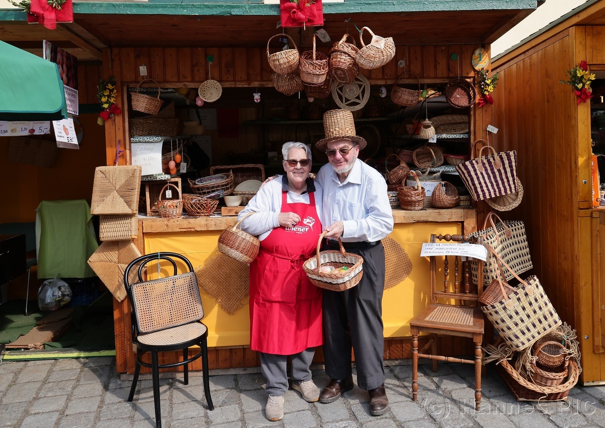 Two people, wearing aprons and hats, stand smiling in front of a wooden stall displaying various handmade baskets.