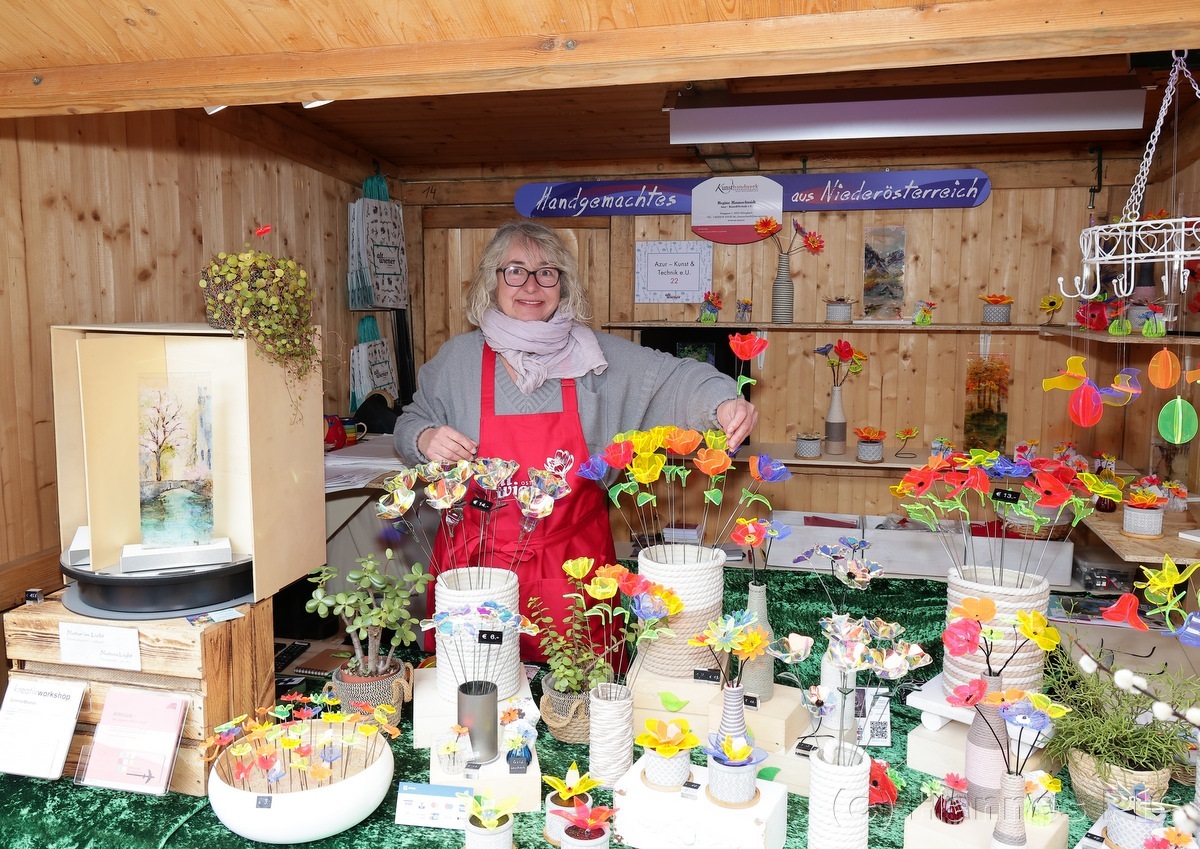 A smiling vendor in a cozy wooden stall displays handmade decorative items and colorful paper crafts on a green tablecloth.