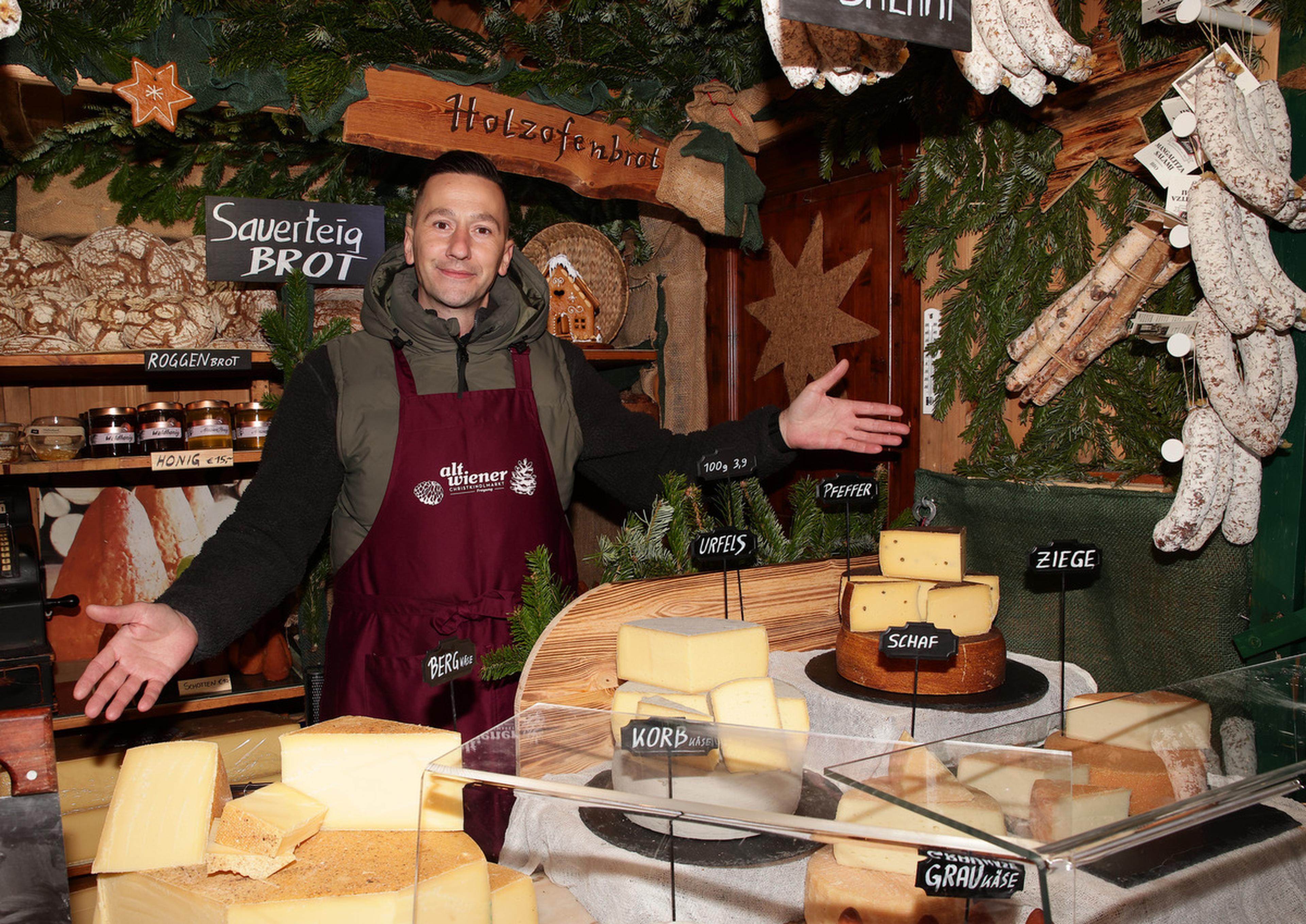 A vendor in a festive market stall displays various cheeses and breads, with hanging sausages and evergreen decorations in the background.