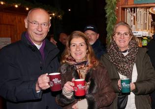 Three people smiling and standing close together at a festive market, holding mugs labeled "alt wien." Warm lighting and wooden stalls in the background.