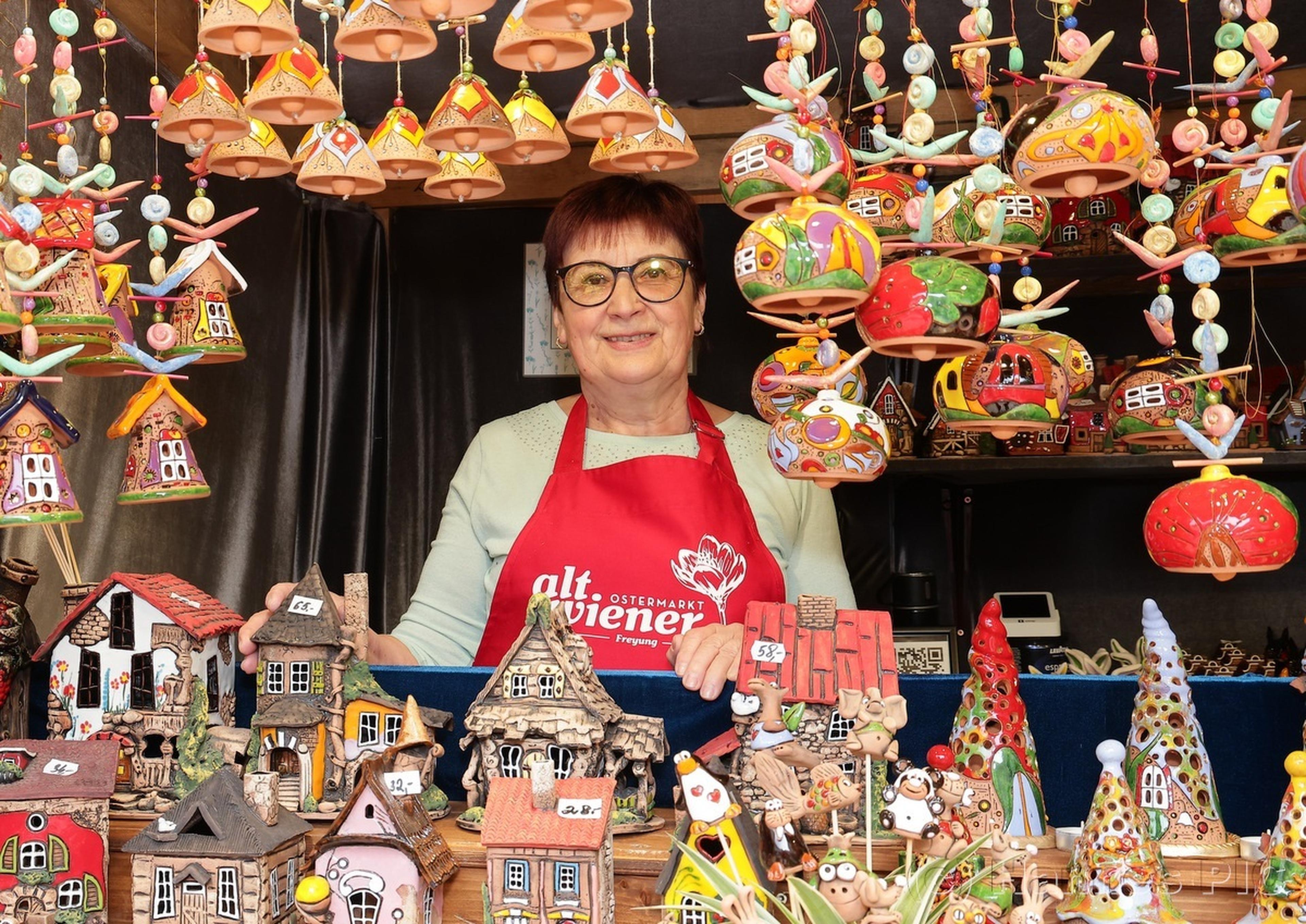 A woman in an apron smiles at a festive market stall displaying colorful clay houses and ornaments.