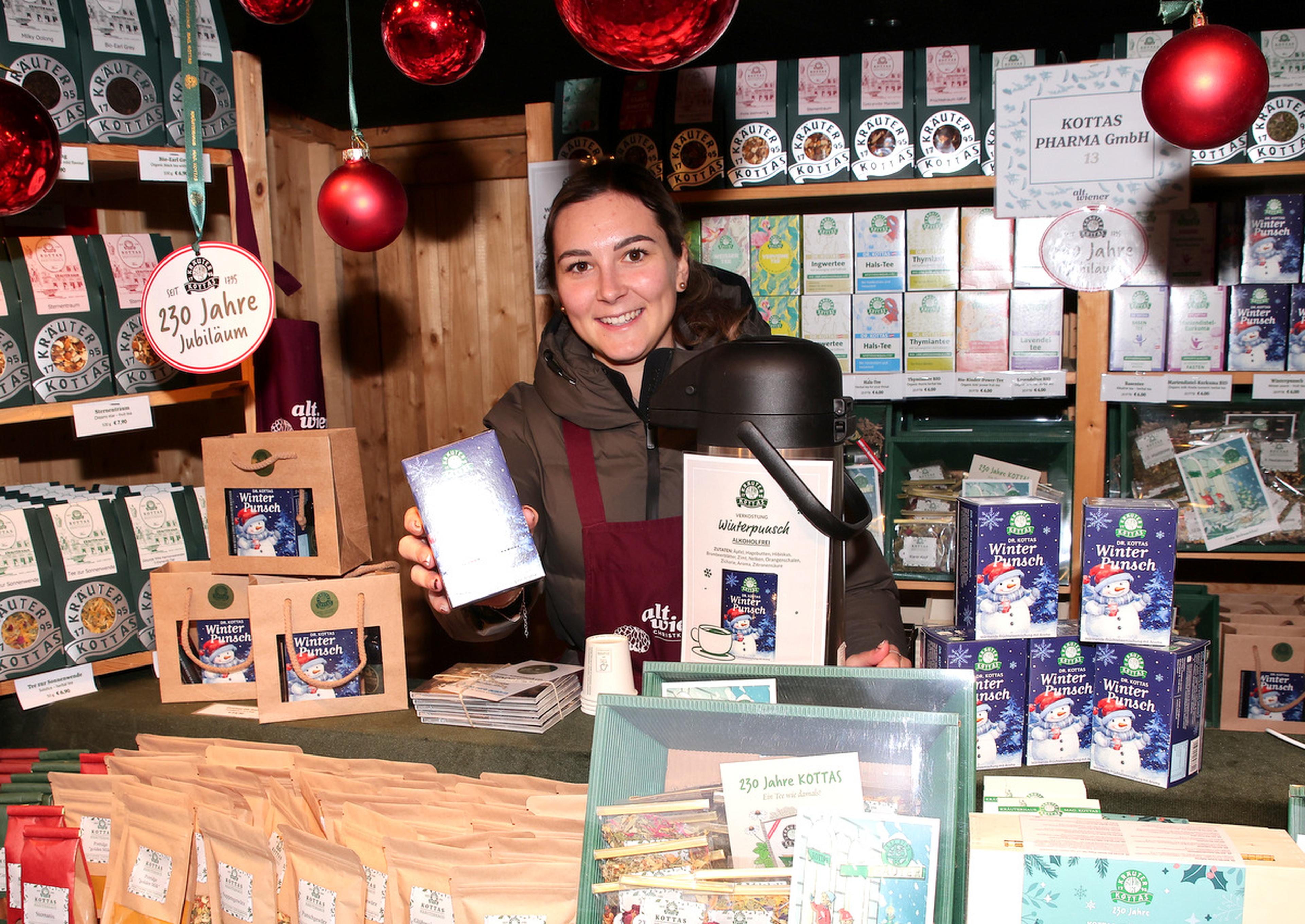 A vendor smiles while holding a tea box at a festive market stall, surrounded by various tea products and holiday decorations.