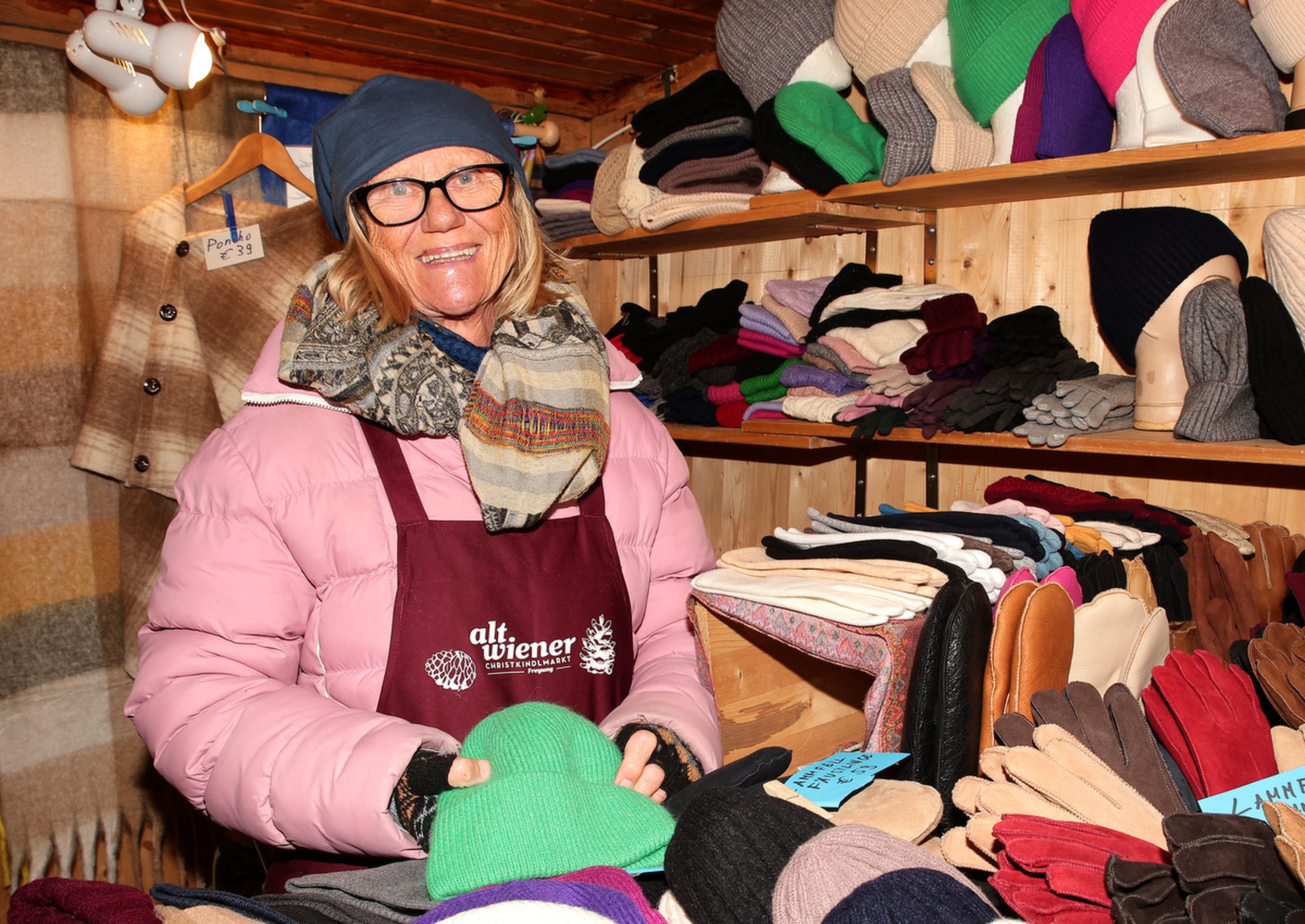 A woman in a pink coat and glasses stands in a market stall, surrounded by colorful hats and gloves, smiling at the camera.