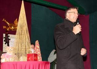 A person in a black coat speaks into a microphone beside a table with decorative trees and gifts on a red cloth.