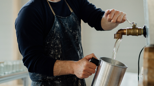 Person filling a metal pitcher with water from a brass faucet, wearing a dark shirt and a paint-splattered apron.