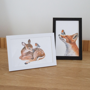 Two framed illustrations on a wooden surface: a fawn with a bird and a fox with a bird. One frame is white, the other is black.