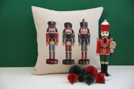 A decorative pillow featuring three embroidered nutcrackers next to a red and green nutcracker figurine and paper ornaments on a green background.