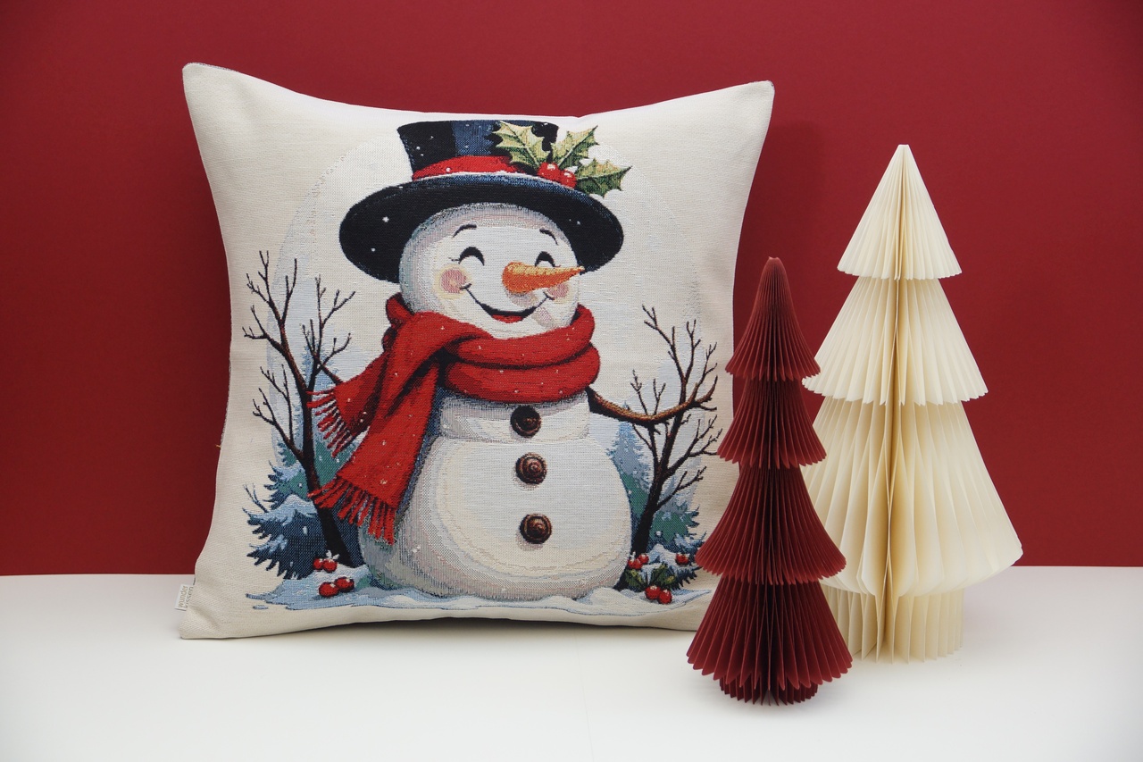 A decorative pillow with a snowman design, wearing a hat and scarf, beside two paper Christmas trees against a red background.
