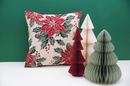 Festive cushion with poinsettia and holly pattern next to three paper Christmas trees on a green background.