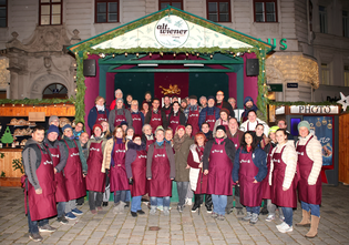 A group of people in matching maroon aprons pose in front of a festive market stall with a sign reading "Alt Wiener."