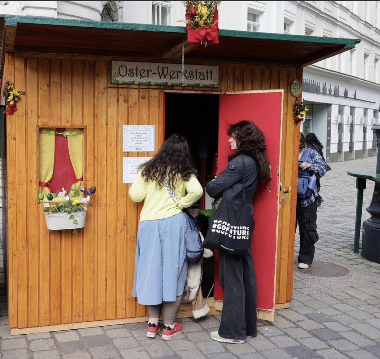 Two women standing at a wooden Easter-themed booth on a cobblestone street, reading posted information. Another person is in the background.