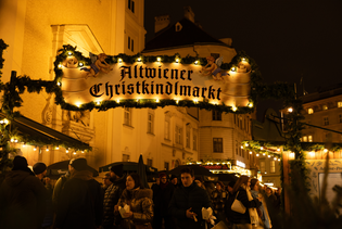People gather at the illuminated entrance of Altwiener Christkindlmarkt, surrounded by festive decorations and warm lighting.