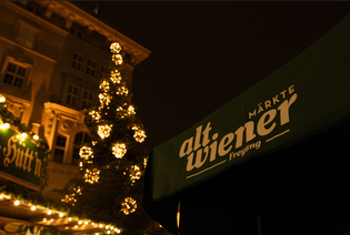 Night scene of a Christmas market with a lit tree and a green awning displaying "alt.wiener märkte" in white text.