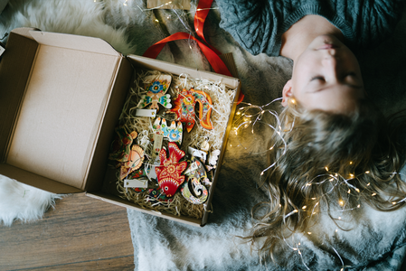 Woman lying on a fuzzy surface beside an open box of colorful ornaments, surrounded by glowing string lights.