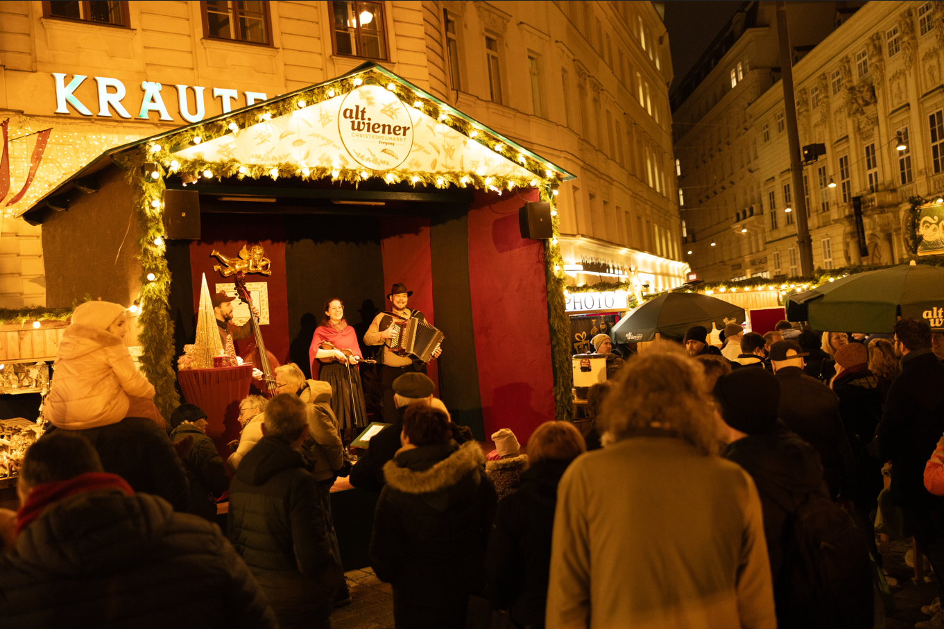 A festive outdoor market scene with musicians performing on a decorated stage, surrounded by a crowd of people on a chilly evening.