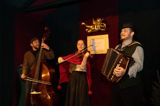 Three musicians perform on stage: one with a double bass, one with a violin, and one with an accordion. A golden cherub decorates the background.