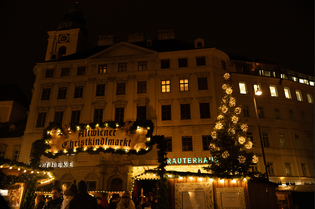 Festively lit Christmas market at night, with a decorated tree and historic building backdrop, featuring a sign for "Altwiener Christkindlmarkt."