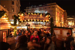 A bustling Christmas market at night with festive lights, a decorative sign reading "Altwiener Christkindlmarkt," and crowds of people.