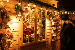 A person in a coat and hat looks at a warmly lit wooden stall filled with colorful Christmas decorations at a festive market.