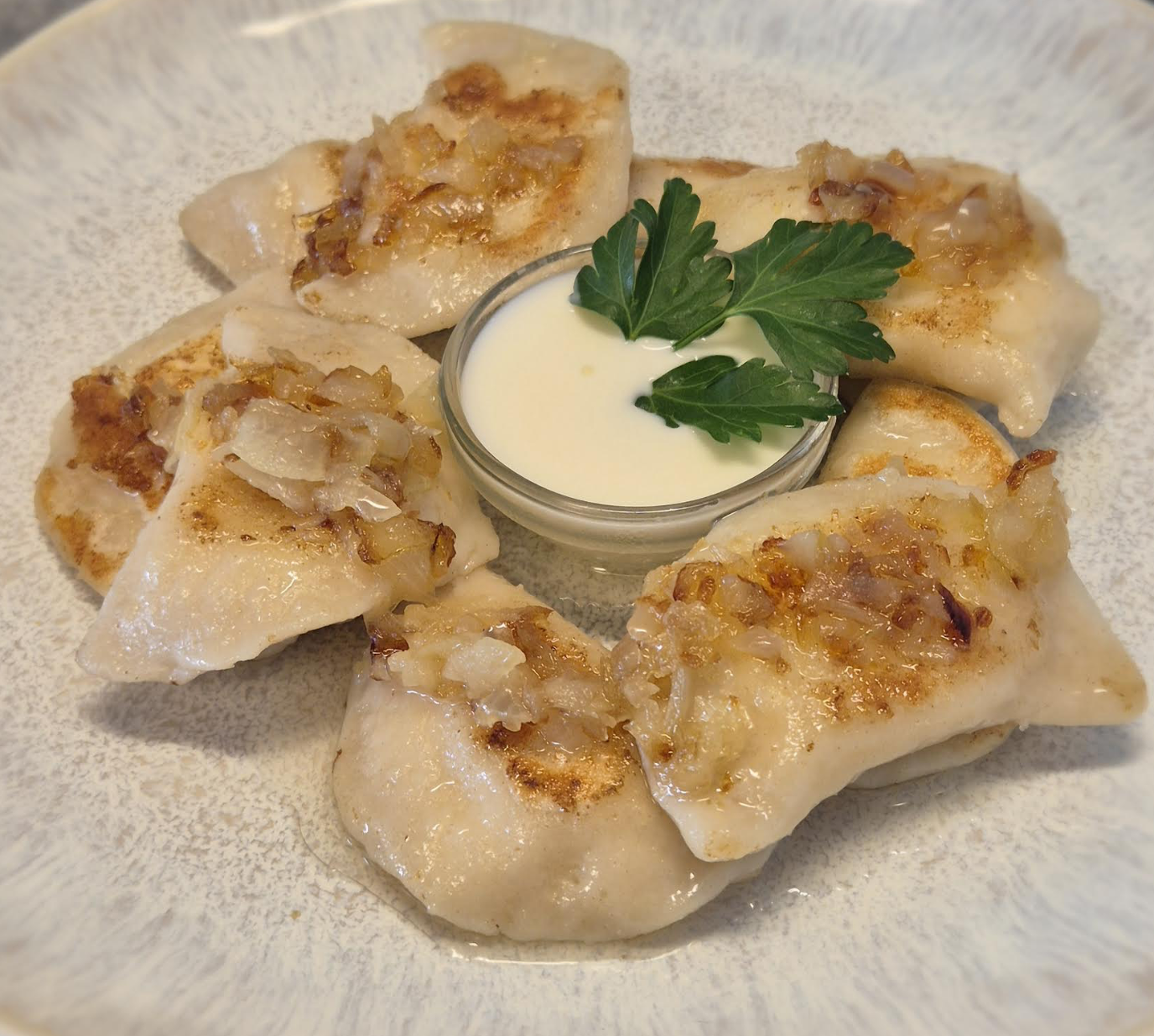 Plate of six golden-brown pierogi garnished with caramelized onions, surrounding a small dish of sour cream with parsley garnish.