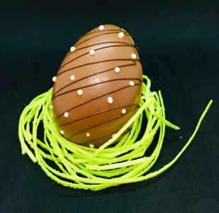 A chocolate egg decorated with white dots and thin dark lines sits on a nest of bright green strings against a black background.