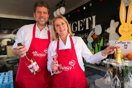 A man and woman in red aprons hold champagne glasses and bottles at a festive stall with Easter decorations.