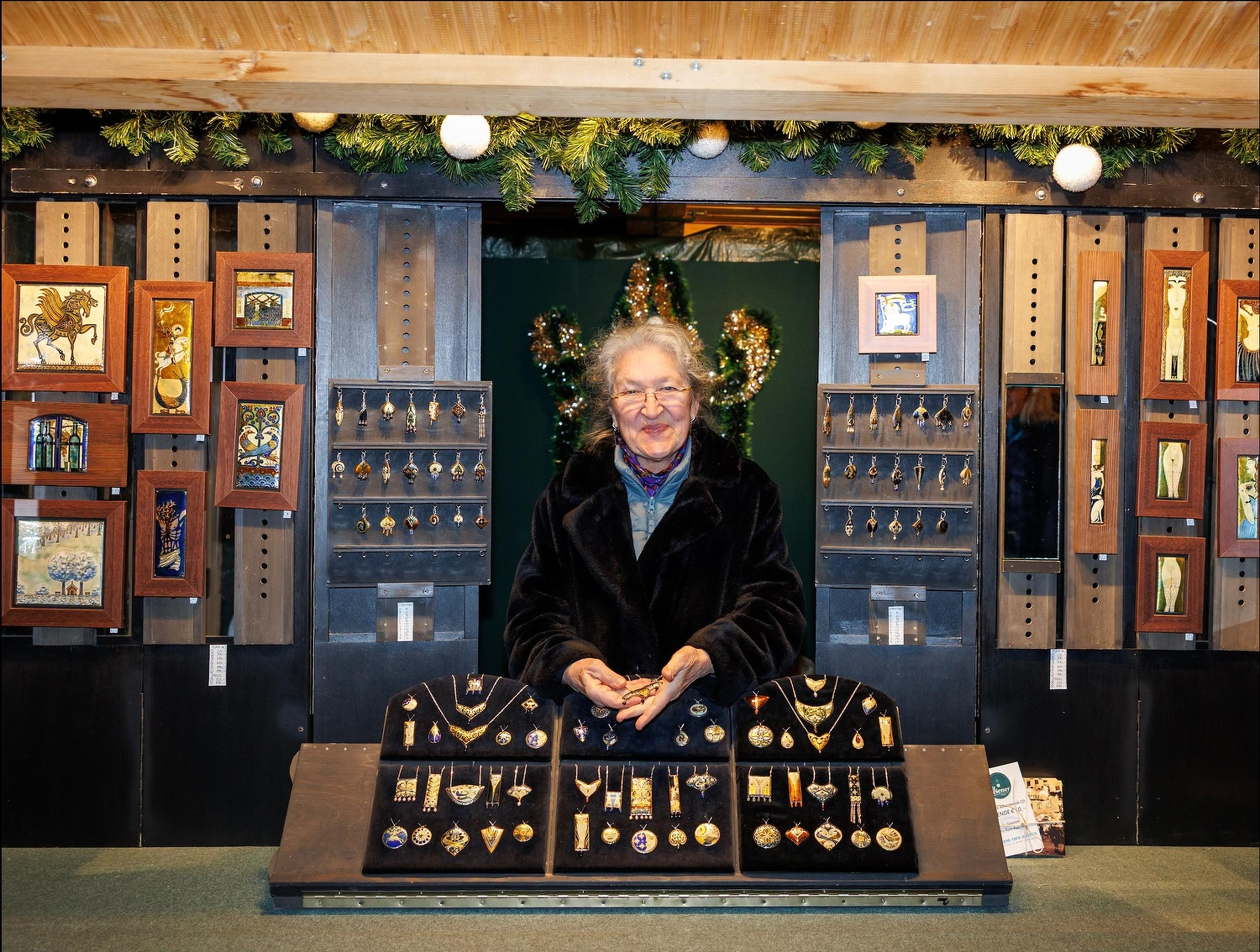 Smiling elderly vendor at a festive market stall displaying handcrafted jewelry and framed artwork beneath pine garland.