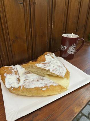 A crispy fried pastry topped with white sauce sits on a paper plate on a wooden table. A red mug filled with a drink is in the background.