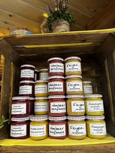 Jars of various mustard sauces with handwritten labels stacked in a wooden crate, set on a yellow cloth.