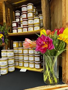 Jars of jam stacked on wooden shelves next to a vase of colorful flowers, including pink, yellow, and red blooms.
