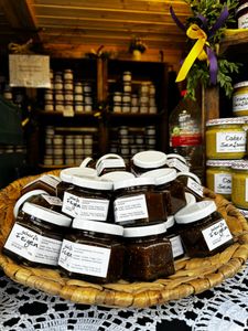 A wicker basket filled with jars of jam, surrounded by various condiments, displayed on a lace-covered table in a rustic shop setting.