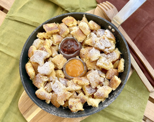 A skillet with Kaiserschmarrn topped with powdered sugar, served with two small bowls of jam, on a wooden and green cloth background.