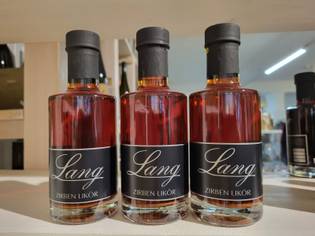 Three bottles of Lang Zirben Liqueur with black labels and gold caps are displayed on a shelf in a well-lit store.