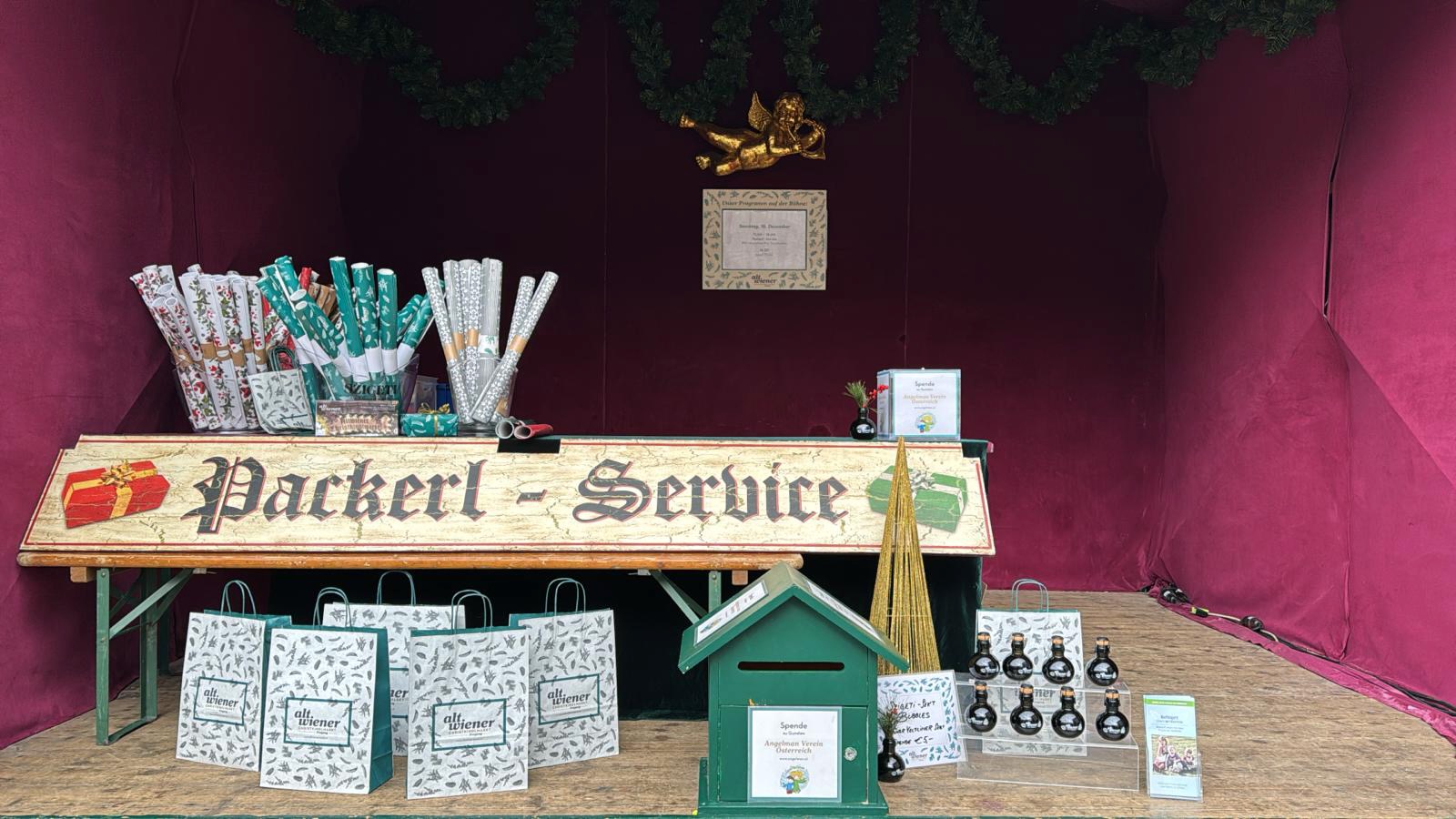 A Christmas-themed wrapping service booth with gift wrap rolls, decorated bags, and bottles on display against a maroon backdrop.