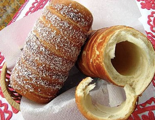 Two chimney pastries on a paper-lined tray; one is coated with sugar and coconut, the other is plain with a bite taken out.