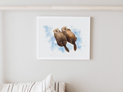 Framed art print of two otters floating on a blue watercolor background, hanging on a white wall above a striped cushion.