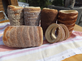Close-up of traditional chimney cakes with various coatings, some whole and some sliced, resting on a striped cloth on a wooden table.